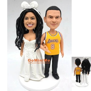 May include: A wedding cake topper featuring a bride and groom. The bride is wearing a white dress and a white bow in her hair. The groom is wearing a yellow basketball jersey with the number 8 on it. The couple is holding hands and smiling.