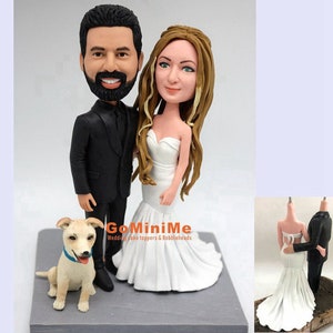 May include: A custom-made wedding cake topper featuring a bride and groom in a white dress and black suit, standing next to a small white dog. The topper is made of sculpted clay and is a personalized gift for a wedding.