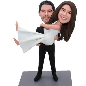 May include: A wedding cake topper featuring a bride and groom figurine. The groom is holding the bride in his arms. The bride is wearing a white dress and the groom is wearing a black tuxedo. The base of the topper is gray and has the text "Mr. & Mrs. Hollinger" written on it.