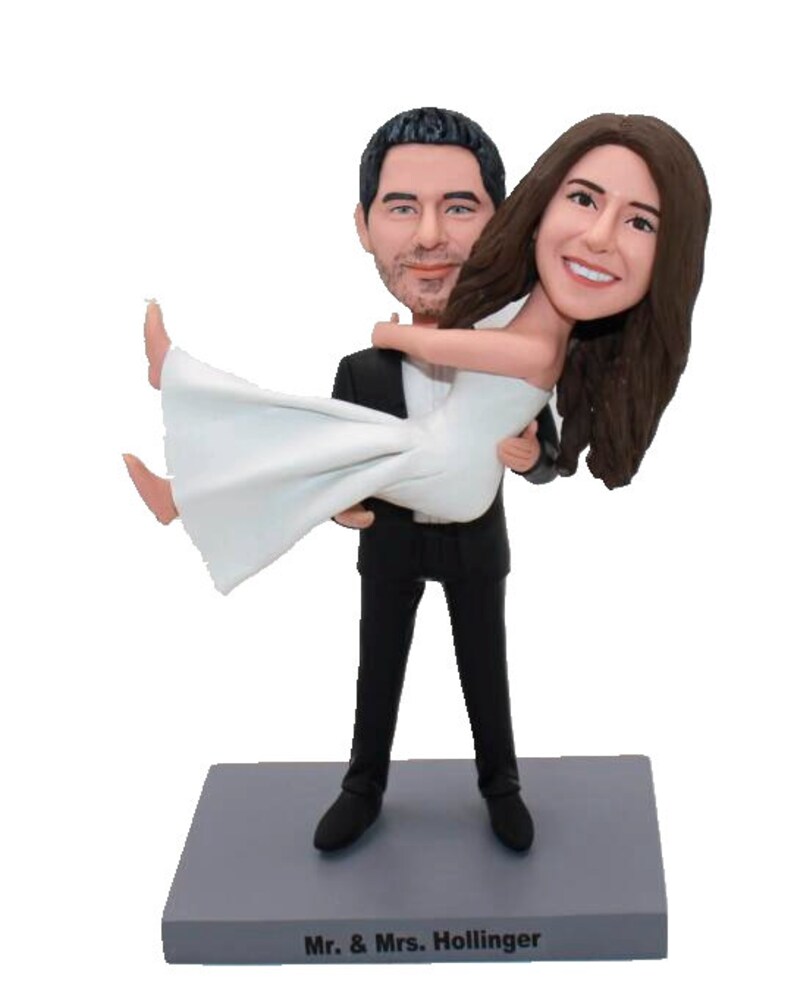 May include: A wedding cake topper featuring a bride and groom figurine. The groom is holding the bride in his arms. The bride is wearing a white dress and the groom is wearing a black tuxedo. The base of the topper is gray and has the text "Mr. & Mrs. Hollinger" written on it.
