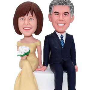 May include: A wedding cake topper featuring a bride and groom in a seated pose. The bride is wearing a yellow dress and holding a bouquet of white flowers. The groom is wearing a dark blue suit and a blue tie.