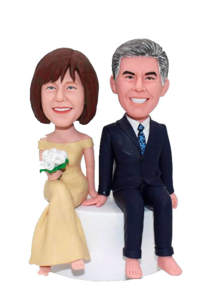 May include: A wedding cake topper featuring a bride and groom in a seated pose. The bride is wearing a yellow dress and holding a bouquet of white flowers. The groom is wearing a dark blue suit and a blue tie.
