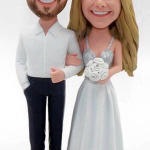 May include: A wedding cake topper featuring a bride and groom in a custom caricature style. The bride is wearing a white dress with a silver sequined bodice and a bouquet of white roses. The groom is wearing a white shirt and black pants. The base of the topper is white with the text "Mr. and Mrs. French 10-19-2019".