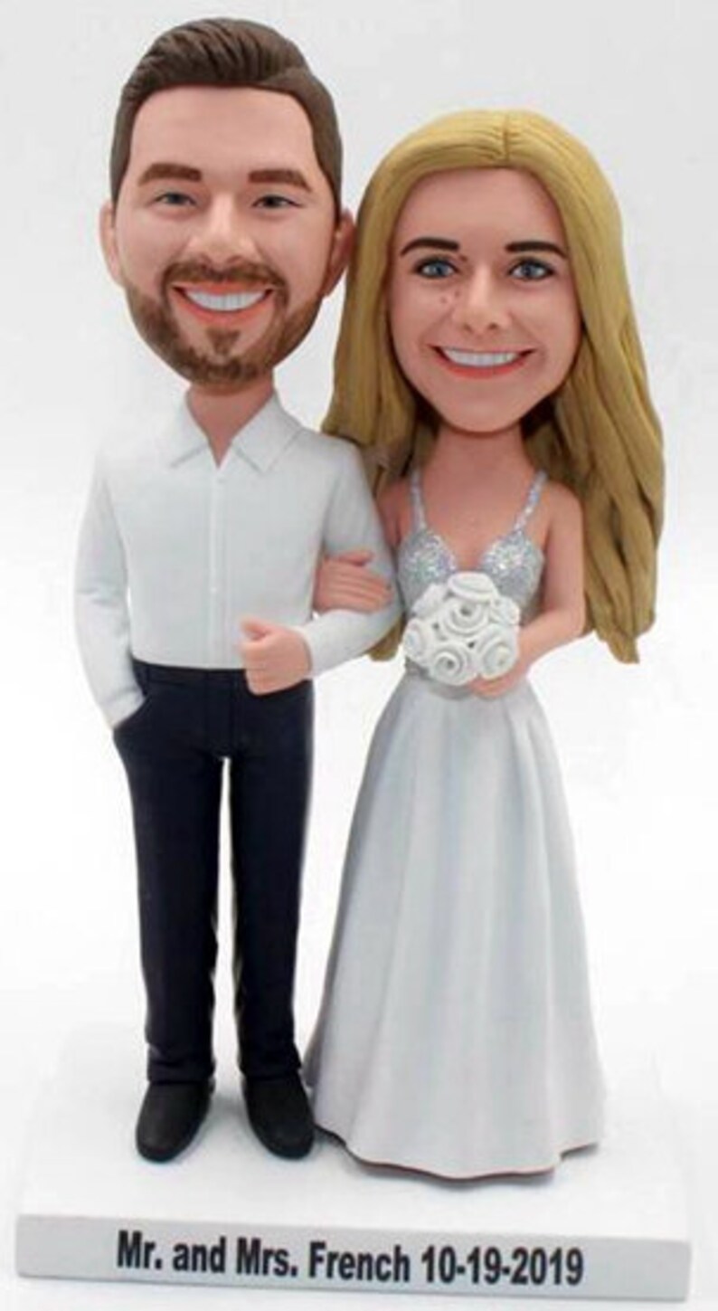 May include: A wedding cake topper featuring a bride and groom in a custom caricature style. The bride is wearing a white dress with a silver sequined bodice and a bouquet of white roses. The groom is wearing a white shirt and black pants. The base of the topper is white with the text "Mr. and Mrs. French 10-19-2019".