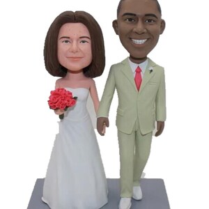 May include: A wedding cake topper featuring a bride and groom in a white dress and a light green suit. The bride is holding a bouquet of red roses. The base of the topper is gray and has the text "Amanda & Eric" written on it.