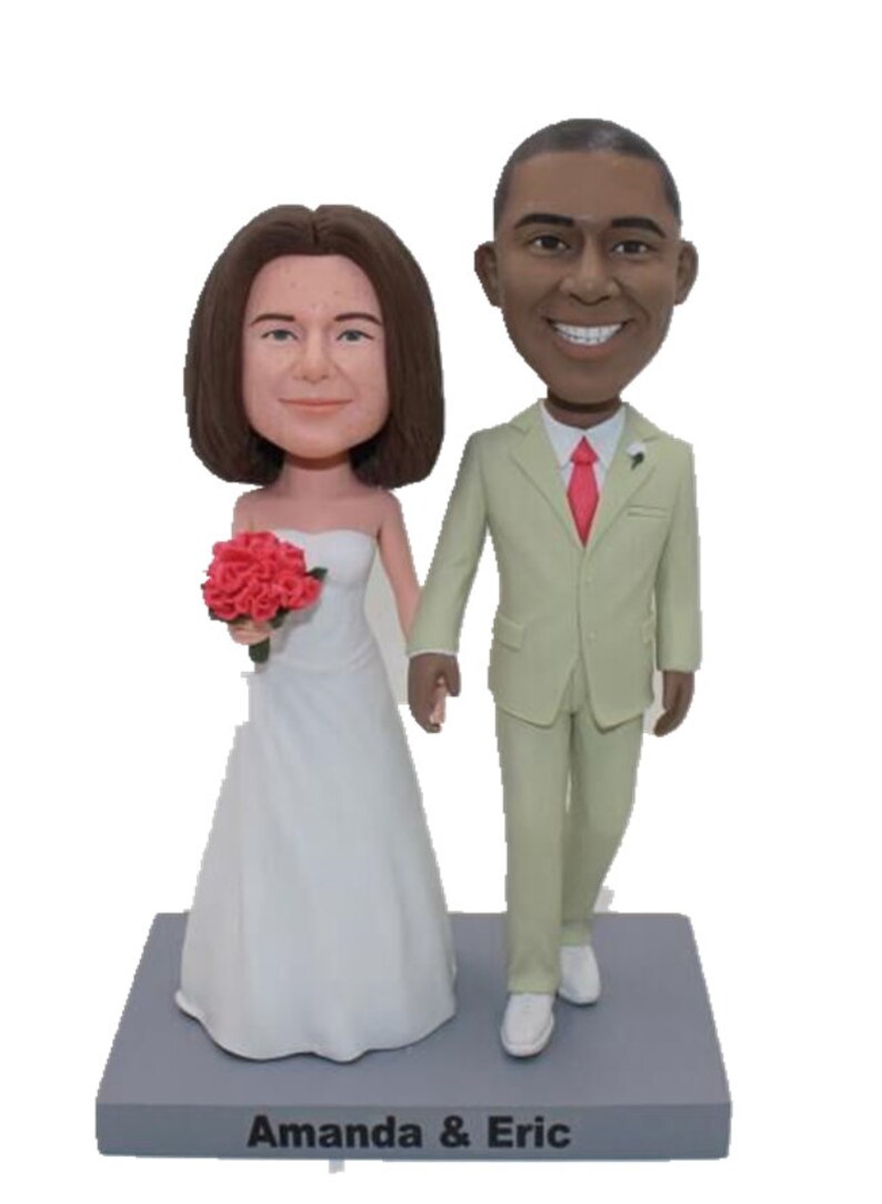 May include: A wedding cake topper featuring a bride and groom in a white dress and a light green suit. The bride is holding a bouquet of red roses. The base of the topper is gray and has the text "Amanda & Eric" written on it.