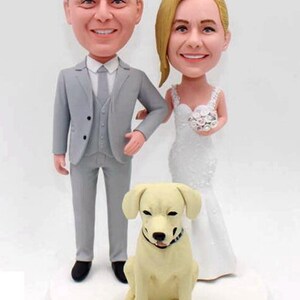 May include: A wedding cake topper featuring a bride and groom in a grey suit and white dress, with a white dog sitting in front of them. The cake topper is on a white base with the date "August 29, 2020" written on it.