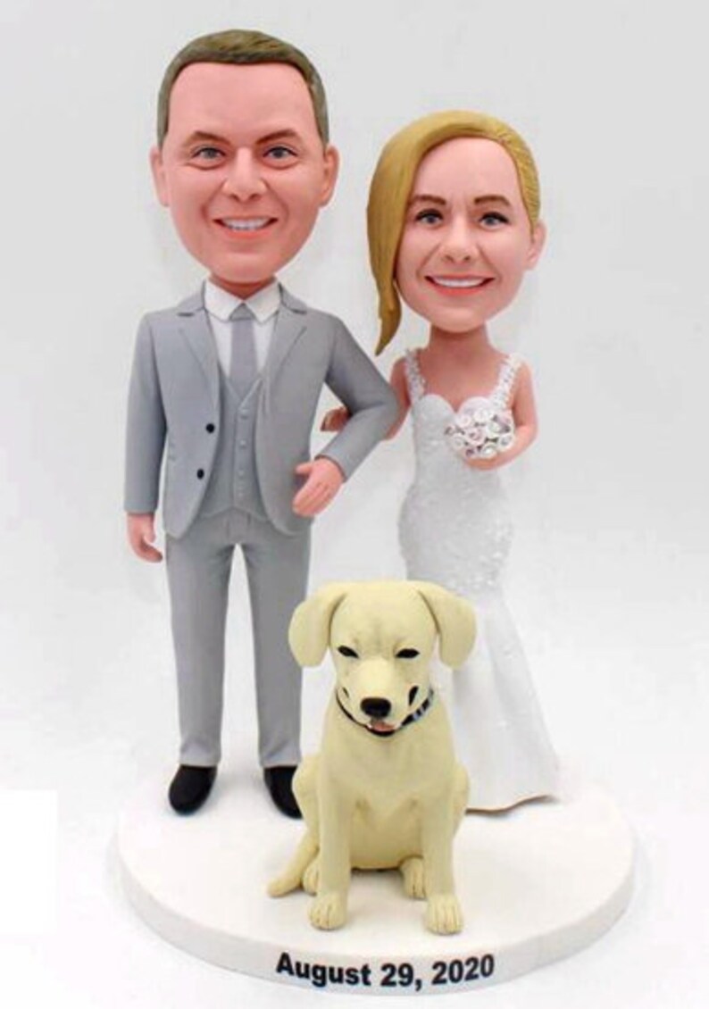May include: A wedding cake topper featuring a bride and groom in a grey suit and white dress, with a white dog sitting in front of them. The cake topper is on a white base with the date "August 29, 2020" written on it.