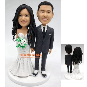 May include: A wedding cake topper featuring a bride and groom in a custom-made figurine style. The bride is wearing a white wedding dress with a lace bodice and a bouquet of white flowers. The groom is wearing a black suit with a white shirt and a black tie. The couple is standing on a white cake base. The text "GoMiniMe Wedding cake toppers & edible heads" is visible on the base.
