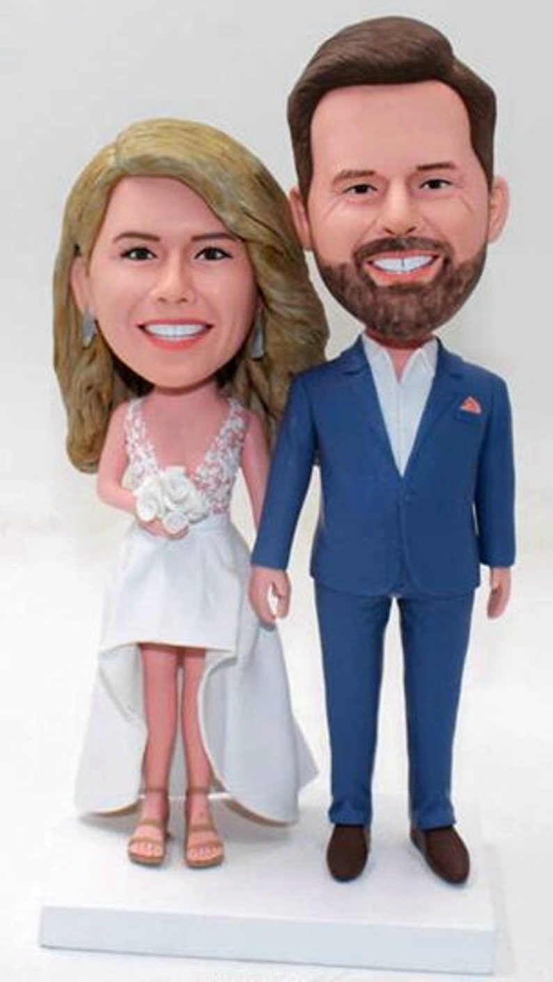 May include: A wedding cake topper featuring a bride and groom in a caricature style. The bride is wearing a white dress with a lace bodice and a bouquet of white flowers. The groom is wearing a blue suit with a white shirt and a red pocket square.