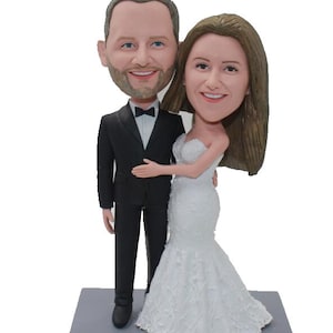 May include: A wedding cake topper featuring a bride and groom in a custom caricature style. The groom is wearing a black tuxedo with a bow tie and the bride is wearing a white wedding dress. The base of the topper is gray and has the text "Tim & Liz June 29th 2019" inscribed on it.