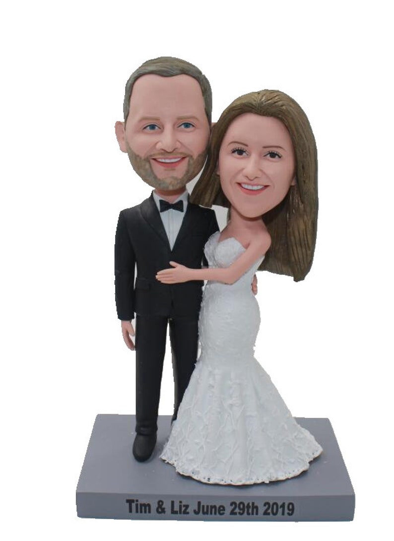 May include: A wedding cake topper featuring a bride and groom in a custom caricature style. The groom is wearing a black tuxedo with a bow tie and the bride is wearing a white wedding dress. The base of the topper is gray and has the text "Tim & Liz June 29th 2019" inscribed on it.