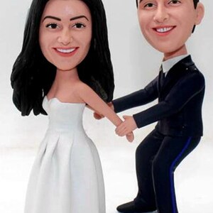 May include: A wedding cake topper featuring a bride and groom in a whimsical, cartoon style. The bride is wearing a white dress and the groom is wearing a dark suit. They are holding hands and smiling.