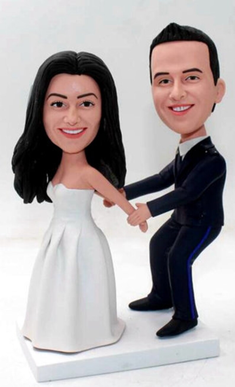 May include: A wedding cake topper featuring a bride and groom in a whimsical, cartoon style. The bride is wearing a white dress and the groom is wearing a dark suit. They are holding hands and smiling.