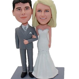 May include: A wedding cake topper featuring a bride and groom in a gray suit and white wedding dress. The base of the topper is gray and has the text "July 6th, 2019" inscribed on it.