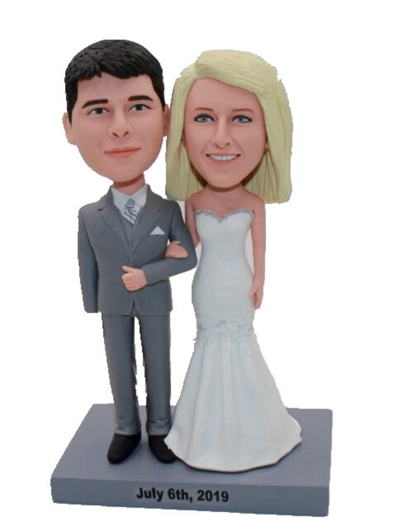 May include: A wedding cake topper featuring a bride and groom in a gray suit and white wedding dress. The base of the topper is gray and has the text "July 6th, 2019" inscribed on it.