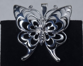 May include: A silver butterfly pendant with intricate cutouts and a detailed design. The pendant is on a black velvet background.