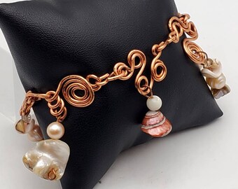 Può includere: Un braccialetto in filo di rame con un design a spirale e accenti di conchiglia. Il braccialetto è esposto su un cuscino nero.