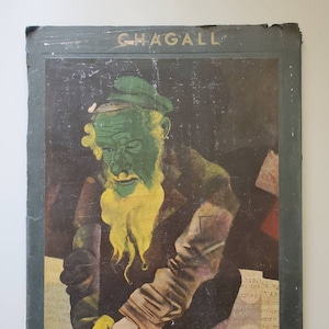 May include: A vintage book cover featuring a painting of a man with a green face and yellow beard. The title of the book is "CHAGALL" and the bottom of the cover reads "SKIRA COLOR PRINTS".