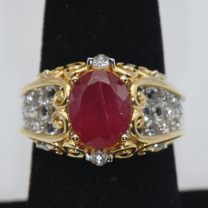 May include: A gold ring with a large oval ruby surrounded by smaller diamonds. The ring has a detailed, ornate design.