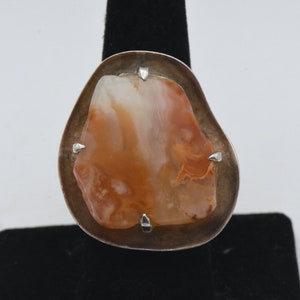 Könnte beinhalten: Ein silberner Ring mit einem großen, unregelmäßig geformten orangefarbenen und weißen Achat-Stein, der in der Mitte eingesetzt ist. Der Stein wird von vier kleinen silbernen Zinken gehalten.