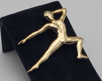 Puede incluir: Broche de metal dorado con forma de figura humana estilizada en una pose dinámica. La figura tiene los brazos extendidos y una pierna doblada, sugiriendo movimiento. El broche se exhibe sobre una superficie de terciopelo negro.