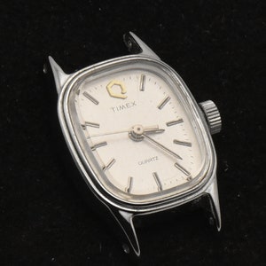 May include: Silver Timex Quartz watch with a rectangular face. The watch has a white dial with black hour markers and hands. The word "TIMEX" is printed above the center, and "QUARTZ" is printed below. The watch is missing its strap.
