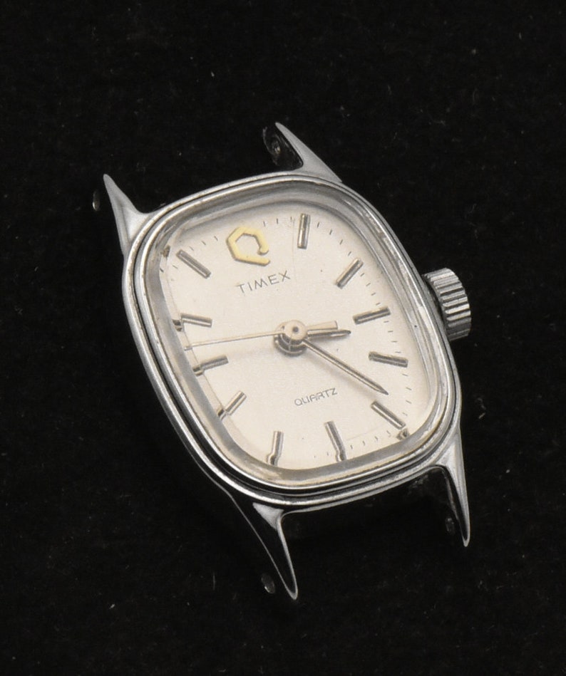 May include: Silver Timex Quartz watch with a rectangular face. The watch has a white dial with black hour markers and hands. The word "TIMEX" is printed above the center, and "QUARTZ" is printed below. The watch is missing its strap.