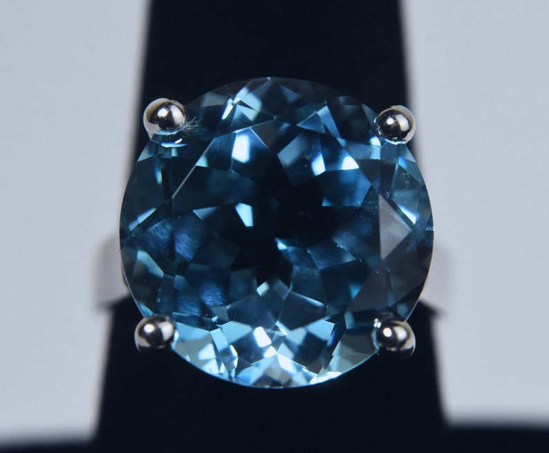 K&ouml;nnte beinhalten: Ein runder blauer Topas-Edelsteinring, der in eine silberne Fassung gefasst ist. Der Ring hat vier Krappen, die den Stein an Ort und Stelle halten.
