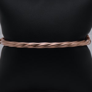 Puede incluir: Un brazalete de oro rosa trenzado. El brazalete está hecho de un metal liso y brillante y tiene un diseño simple y elegante.