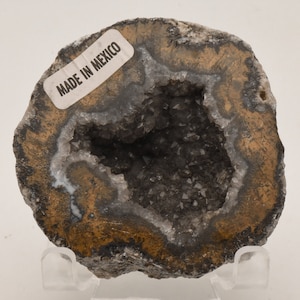 May include: A geode with a brown, gray, and white exterior. The geode is open, revealing a cavity lined with small, clear crystals. The geode has a label that reads "Made in Mexico".