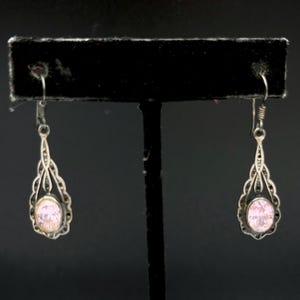 May include: A pair of silver drop earrings featuring an oval-shaped light pink stone set within a decorative filigree frame. The earrings hang from simple silver hooks and exhibit a detailed metalwork design surrounding the central gemstone.