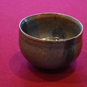 Puede incluir: Un cuenco de cerámica marrón con un esmalte moteado. El cuenco tiene un borde marrón oscuro y un interior marrón claro.