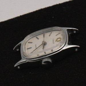 May include: Silver Timex Quartz watch with a light-coloured face. The watch has a rectangular shape with curved edges and a small crown. The watch face displays the word "TIMEX" and "QUARTZ".