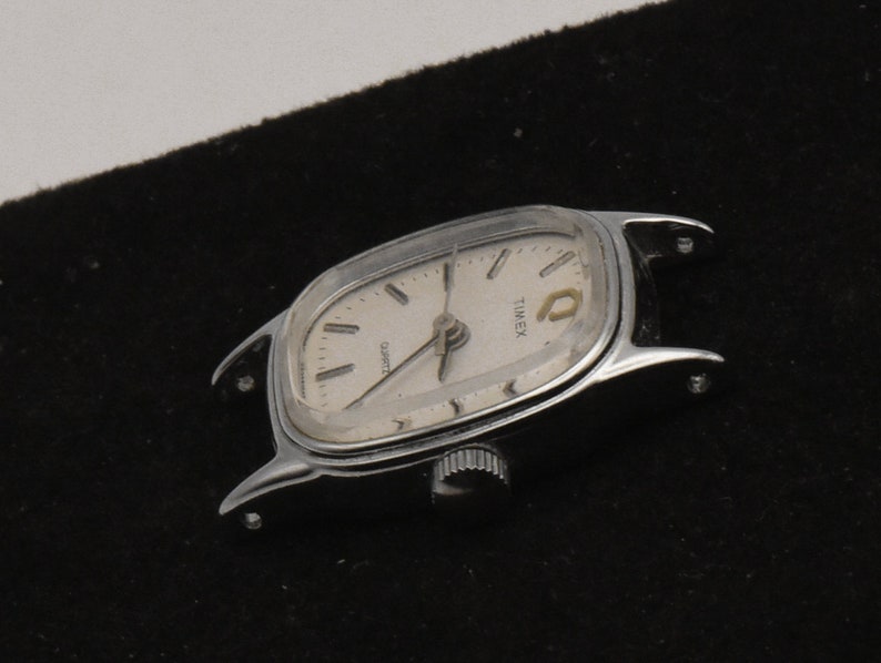 May include: Silver Timex Quartz watch with a light-coloured face. The watch has a rectangular shape with curved edges and a small crown. The watch face displays the word "TIMEX" and "QUARTZ".