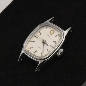 May include: A silver Timex Quartz watch face with a rectangular shape and a cream dial. The watch has a small crown and the word "TIMEX" is printed on the face. The watch case has extended lugs.
