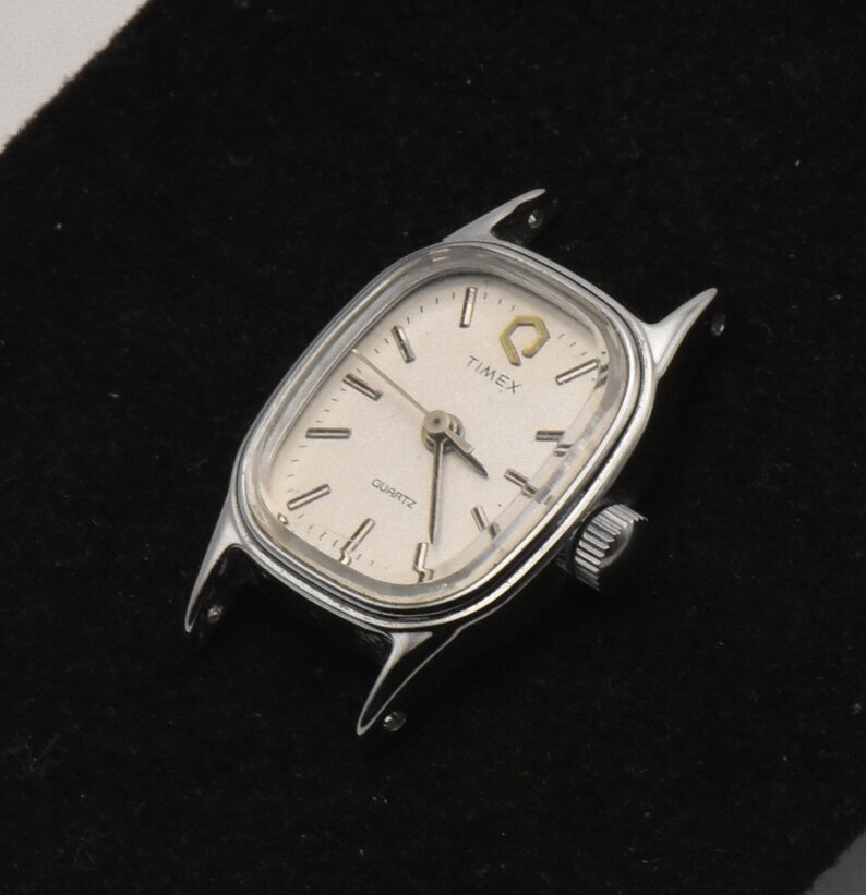 May include: A silver Timex Quartz watch face with a rectangular shape and a cream dial. The watch has a small crown and the word "TIMEX" is printed on the face. The watch case has extended lugs.