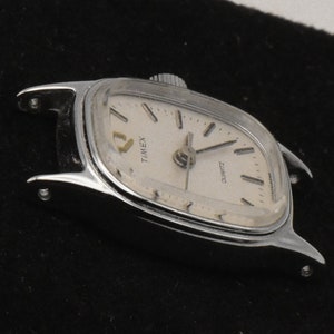 May include: A vintage Timex Quartz watch with a silver case and a cream face. The watch has a rectangular shape with curved edges and a small crown. The watch face displays the brand name and the word "QUARTZ".