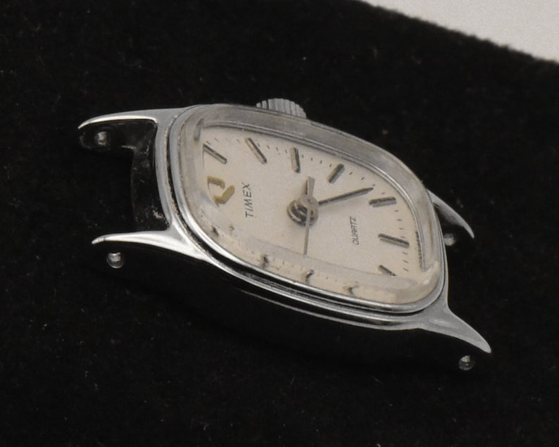 May include: A vintage Timex Quartz watch with a silver case and a cream face. The watch has a rectangular shape with curved edges and a small crown. The watch face displays the brand name and the word "QUARTZ".