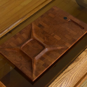 Puede incluir: Una bandeja de servir rectangular de madera con tres compartimentos separados. La bandeja es de madera oscura con un patrón de cuadros y tiene una pequeña manija negra.