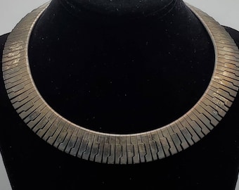Puede incluir: Un collar gargantilla de tono plateado con un diseño geométrico. El collar está hecho de piezas de metal entrelazadas que crean un aspecto único y moderno.
