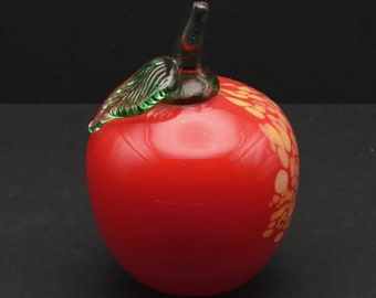 May include: A red glass apple with a green glass stem and leaf. The apple has a speckled pattern of yellow and white.