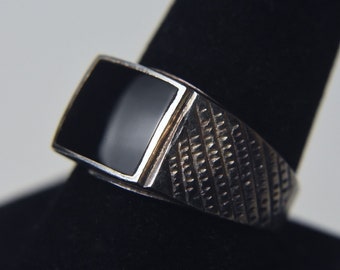 May include: A silver ring with a black square stone set in the center. The band of the ring has a textured design.