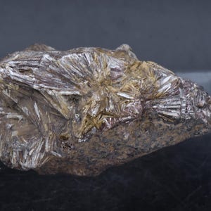 Puede incluir: Un espécimen mineral en bruto, de forma irregular y con una mezcla de colores. La piedra presenta una matriz de tonos marrones y grises, con estructuras cristalinas radiantes en tonos plateados y dorados. La textura general es áspera y natural.