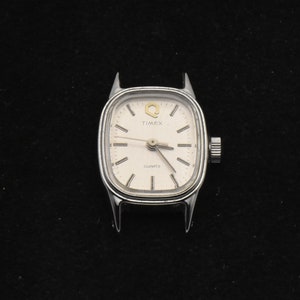 May include: A silver Timex Quartz watch face with a white dial. The watch has a rectangular shape with rounded corners and a crown on the right. The watch face displays the Timex logo and the word "QUARTZ".