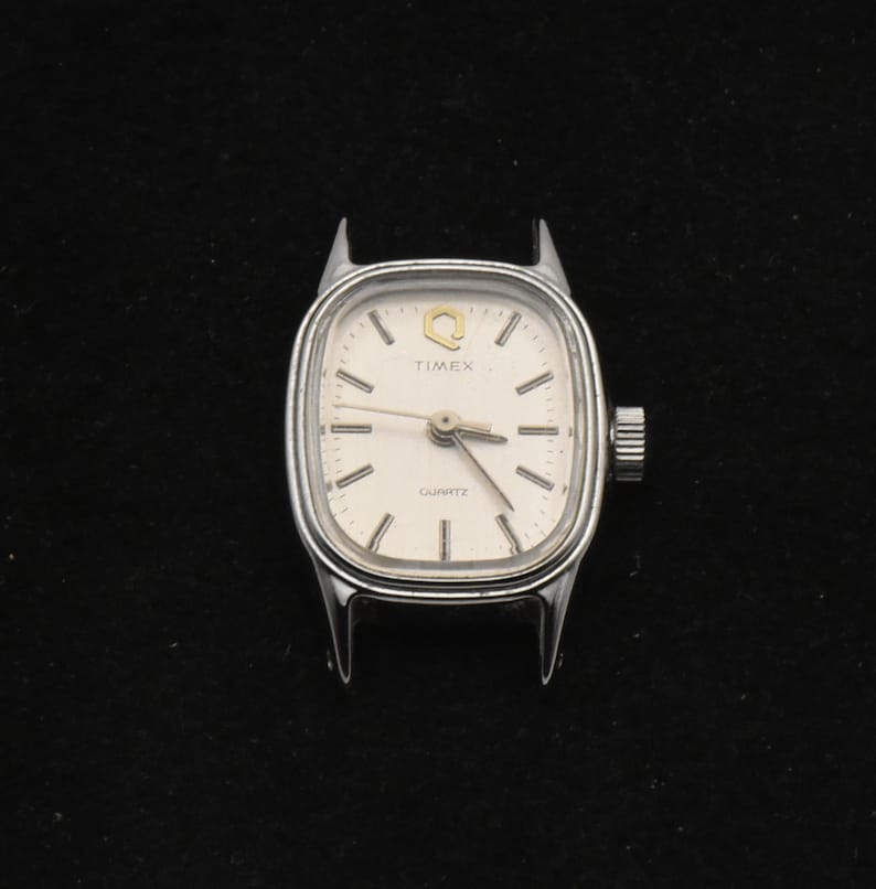 May include: A silver Timex Quartz watch face with a white dial. The watch has a rectangular shape with rounded corners and a crown on the right. The watch face displays the Timex logo and the word "QUARTZ".