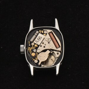 May include: A silver watch case, back removed, showcasing the internal mechanisms. The watch face reveals intricate components, including a coil, gears, and the text "A66". The watch features a crown for winding and setting the time.