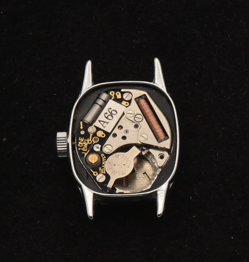 May include: A silver watch case, back removed, showcasing the internal mechanisms. The watch face reveals intricate components, including a coil, gears, and the text "A66". The watch features a crown for winding and setting the time.