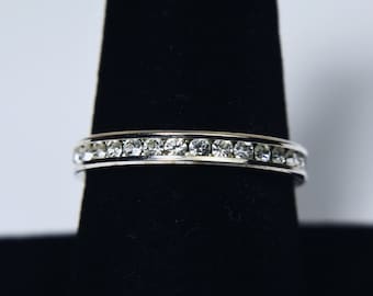 May include: A silver ring featuring a continuous row of clear gemstones. The ring is displayed on a black stand, highlighting its design. The gemstones are evenly set around the band, creating a sparkling effect. A piece of jewellery.