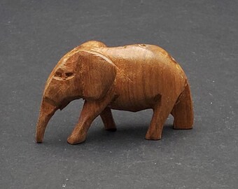 May include: A small, hand-carved wooden elephant figurine. The elephant is brown with visible wood grain and carved details, including a trunk, ears, and legs. The figurine is standing on a dark surface.